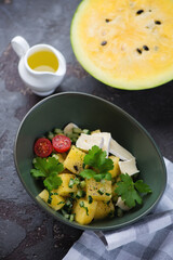 Green bowl with yellow watermelon, herbed cucumber and cheese salad, vertical shot on a brown stone background