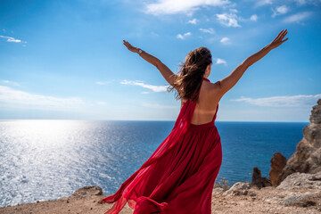 From behind, a woman is seen in a red flying dress fluttering in the wind. In a straw hat, walking down the stairs against the background of the sea with rocks. The concept of travel