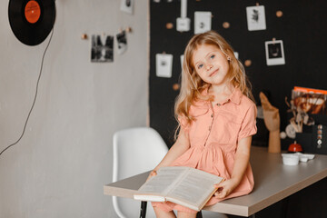 little cute blonde girl with a book sitting on the table in a pink dress in the room