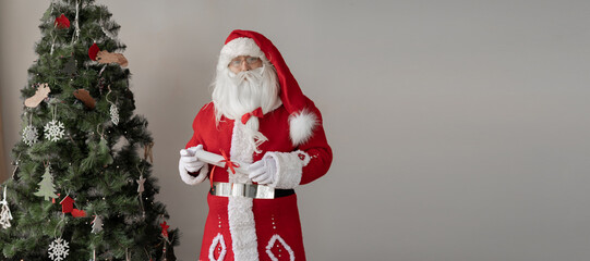 Santa Claus is standing near the Christmas tree, holding a rolled scroll of paper. Red ribbon and bow.