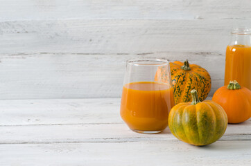 Pumpkin vitamin juice in a glass glass on a white wooden background. Orange little pumpkins lie nearby, copy space, selective focus