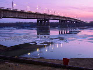 Ice drift on the Irtysh river under the Leningrad bridge at sunset in spring. Lighting is lit on the bridge. The inscription on the plate: the passage (moving) on â€‹â€‹the ice is forbidden.