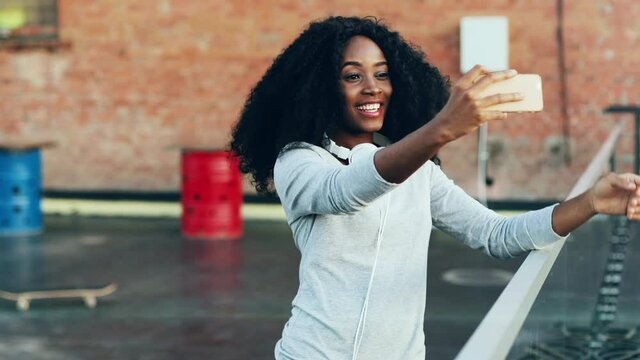 Cheerful young woman with curly hair stands on terrace and recording video on her smartphone. Beautiful african american woman standing on roof talking on video call holding phone in outstretched hand