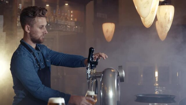 Barkeeper in apron draft glass of beer with foam crown in smoky bar shot in 4k super slow motion 