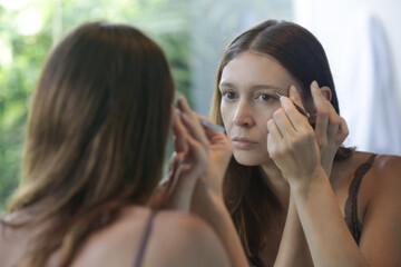 Young caucasian woman plucking her eyebrows in front of the mirror.	