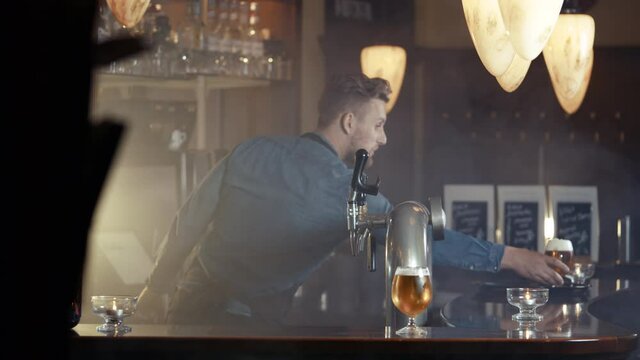 Busy barkeeper in apron draft glass of beer with foam crown in smoky bar shot in 4k