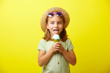 Little caucasian girl in green dress and straw hat, eating beautiful ice cream, wears blue sunglasses, has a good mood, stands on yellow isolated.
