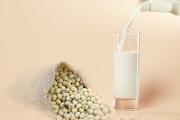 Soy milk in the glass by pouring from bottle with uncooked soybean or soya in sack as pastel orange colors background. Healthy food and vegetarian concept.