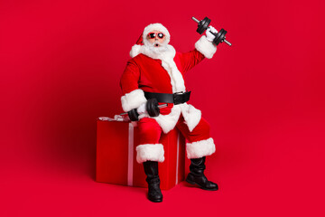 Full length photo of retired old man white beard sit giftbox hold dumbbells shocked easy workout wear x-mas santa costume glove coat belt sunglass cap boot isolated red color background