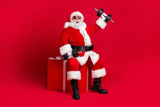 Full Length Photo Of Retired Old Man White Beard Sit Giftbox Hold Dumbbells Shocked Easy Workout Wear X-mas Santa Costume Glove Coat Belt Sunglass Cap Boot Isolated Red Color Background