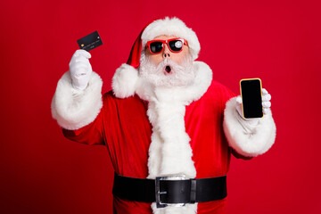 Portrait of nice handsome amazed stunned bearded Santa holding in hand plastic card device web app shop buy order delivery pay wireless payment isolated bright vivid shine vibrant red color background