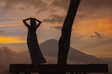 Silhouette of a girl with hands behind head in a long dress stands with Bali landmark volcano Agung view. Travel blogger, Indonesia best sunset point destination. Woman retreat. High quality photo