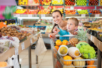 Happy cheerful little boy with his positive smiling mother choosing fresh fruits and vegetables at supermarket