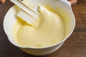 Folding smooth light yellow cake mixture with a spatula in a white bowl on top of wood table background.