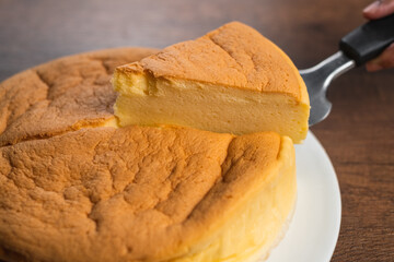 Fluffy baked Japanese cheesecake, lifting a cut piece with a cake server. Cake on white plate on wood tabletop background.