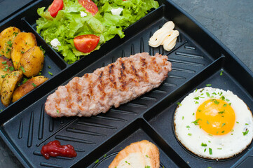 Restaurant lunch, content for the site. Breakfast, cutlet or steak, tomato salad, fried potatoes, croutons and fried egg on black plate.