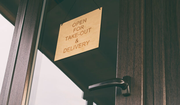 The wooden sign with text: Open for take-out and delivery hanging on the door in cafe