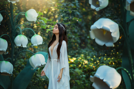 A beautiful elf girl with long hair walks in a fantasy forest. Huge flowers lilies of the valley, green trees, creative decorations. White long medieval dress. Woman inhales smells a scent of flowers.