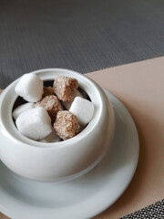 Lots of white and brown sugar cubes in a white ceramic sugar bowl on a white saucer on a table covered with a beige tablecloth and brown napkins
