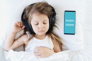 Little girl listening to children's audiobook in mobile phone in headphones before sleeping in bed. Education, child development. E-book with fairy tales, lullaby for the night. Modern technology