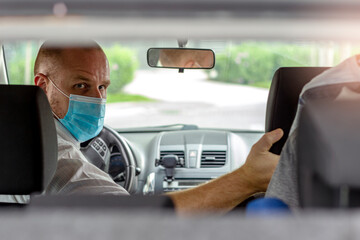 Rear view image of a happy driver wearing a blue protective face mask in the car and looking at the camera,COVID-19 lifestyle concepts.Young taxi driver in medical face mask inside car looks at camera