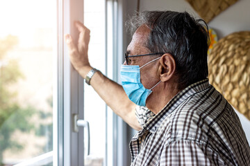 Fototapeta premium Shot of a senior man wearing a respirator or surgical face mask while looking out of the window at home.Elder man with protective face mask looking out of window. Man in home quarantine wearing a mask
