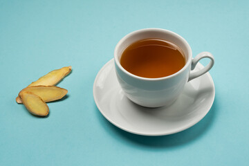 Healthy ginger tea in a white porcelain tea cup with slices of ginger on a cyan clean surface with space for text. 
