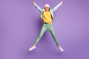 Fototapeta na wymiar Full body photo of funky pretty lady stylish clothes jump high rejoicing spread hands star shape wear sun specs casual hat modern jacket green pants footwear isolated purple color background