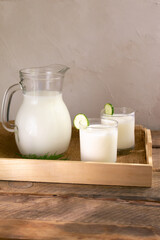 Two glasses with Ayran with a cucumber and a glass jug on a wooden tray on a wooden background. Fermented product concept. Copy space. Vertical orientation.