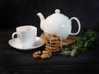 White teapot with a white cup, cookies with chocolate, branches of mint on the dark wooden background. 