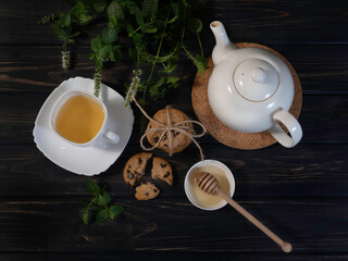 Green tea in the white cup, white teapot, cookies, honey, branches of mint on the dark wooden...