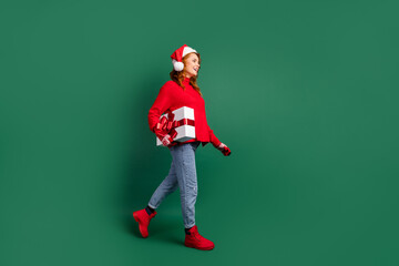 Full length profile photo of funny girl carry present box walk wear red pullover x-mas cap jeans footwear isolated green color background