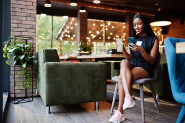 Fashionable feminist african american woman wear in black t-shirt and shorts, posed at restaurant with lemonade glass and mobile phone.