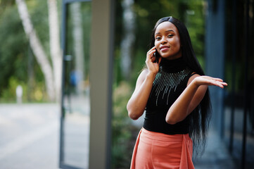 Fototapeta premium Fashionable african american woman in peach pants and black blouse speak on phone outdoor.