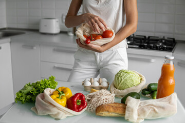 Woman's hand, holding a reusable grocery bag with vegetables on a kitchen at home and takes tomato out. Zero waste and plastic free concept. Mesh cotton shopper with vegetables. Ecology.