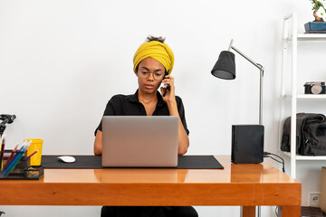Shot of young latin woman working at home office with laptop speaking on mobile phone
