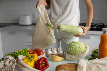 Woman's hand, holding a reusable grocery bag with vegetables on a kitchen at home and takes zucchini out. Zero waste and plastic free concept. Mesh cotton shopper with vegetables. Ecology.