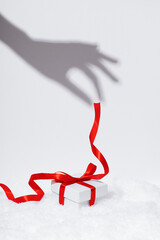 Female hand holding a shadow of a red gift ribbon on a white background. White box for wrapping gifts on a snowy background. Christmas composition with new year decor