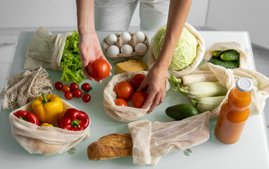 Woman's hand, holding a reusable grocery bag with vegetables on a kitchen at home and takes tomato out. Zero waste and plastic free concept. Mesh cotton shopper with vegetables. Ecology.