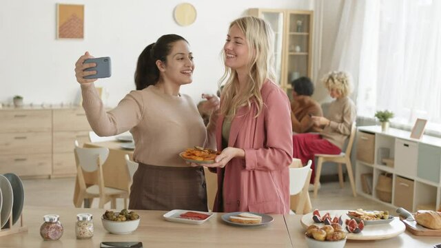 Medium shot of two multiethnic women standing at kitchen table and making selfie on smartphone camera while their friends sitting on background and talking