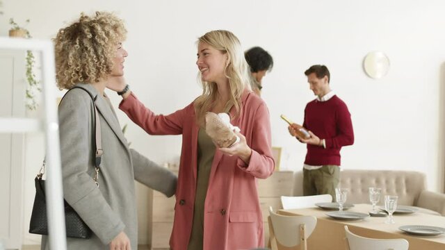 Medium shot of Caucasian curly-haired woman giving present to her female friend and hugging her while two diverse men discussing wine bottle on background