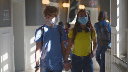 High school boy and girl students in mask walking in corridor holding hands