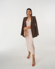 Elegant woman in peach color silk slip dress and brown squared wool blazer standing and posing in...