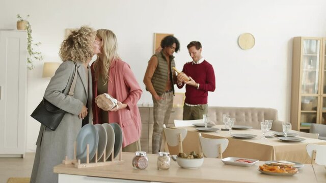 Wide shot of Caucasian blond-haired woman hugging her female friend with curly hair coming and giving her present while two diverse men discussing wine bottle on background