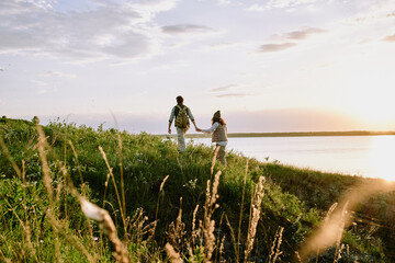 Obraz premium Rear view of young man holding hand of girlfriend while they hiking hills together at sunset