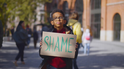Portrait african schoolboy standing near school building and holding cardboard sign with shame text