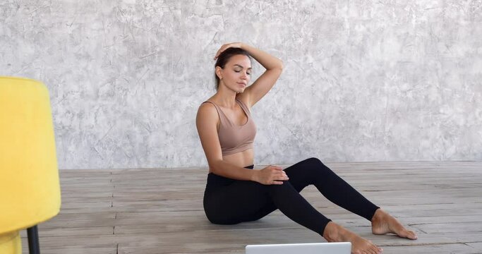 Girl trainer or dancer woman alone at home in room sits on floor near yellow chair, feels pain in neck, does exercises on back, pulls muscles tilting her head to sides, trains online looking at laptop