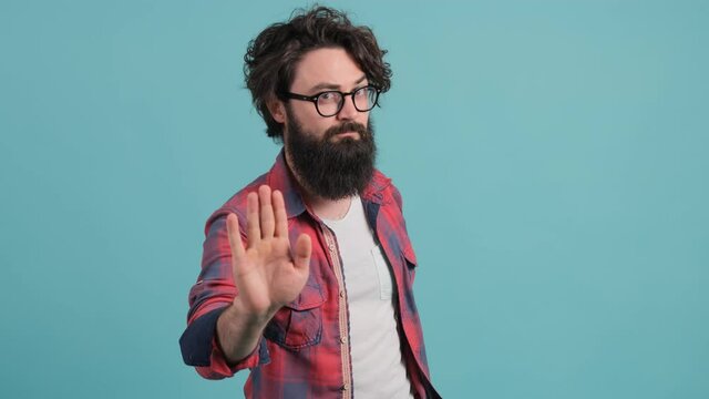 Close up of a young bearded man annoyed with bad attitude making stop sign with hand, saying no, expressing security over turquoise background.