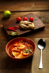 Tom yam soup with shrimp and lime in a brown dish on a wooden table next to a spoon on a napkin and ingredients lime ginger.