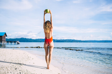 Back view of slim woman holding tropical coconut fruit over head standing at seashore beach, tan...